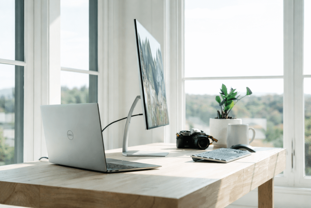 Clean minimalist desk setup with laptop, external monitor, keyboard, camera, and small plant near large bright windows with natural daylight.